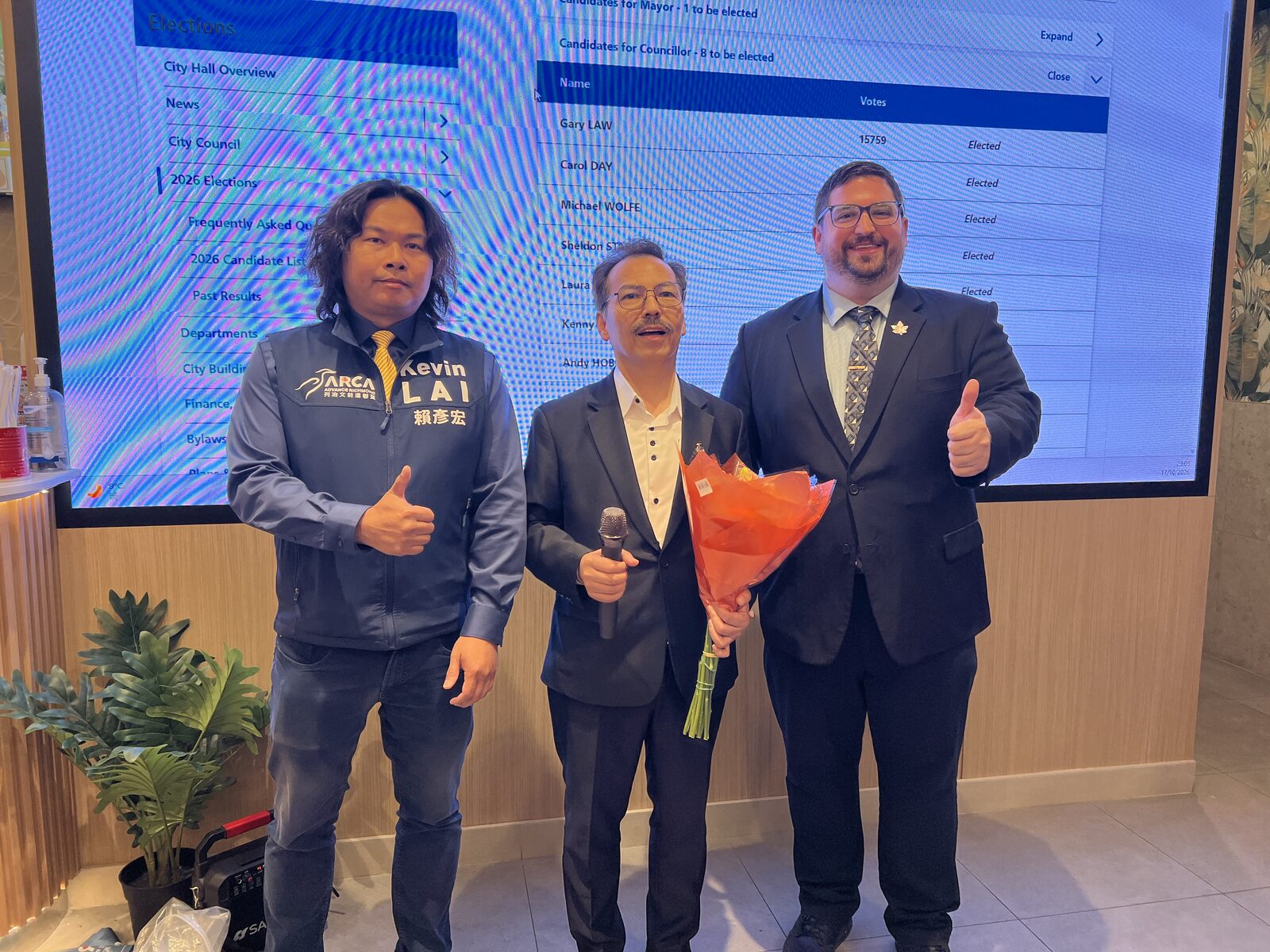 Three people pose with a bouquet and microphone in front of a screen displaying Richmond council election results